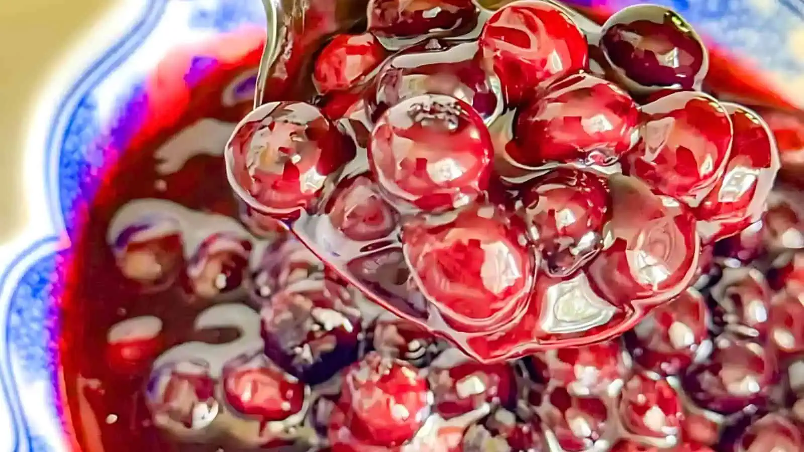 Close-up of a spoon holding several glossy, red cherries with a rich, thick syrup dripping off them. The background shows a bowl filled with more cherries and syrup, with a decorative pattern along the rim of the bowl.