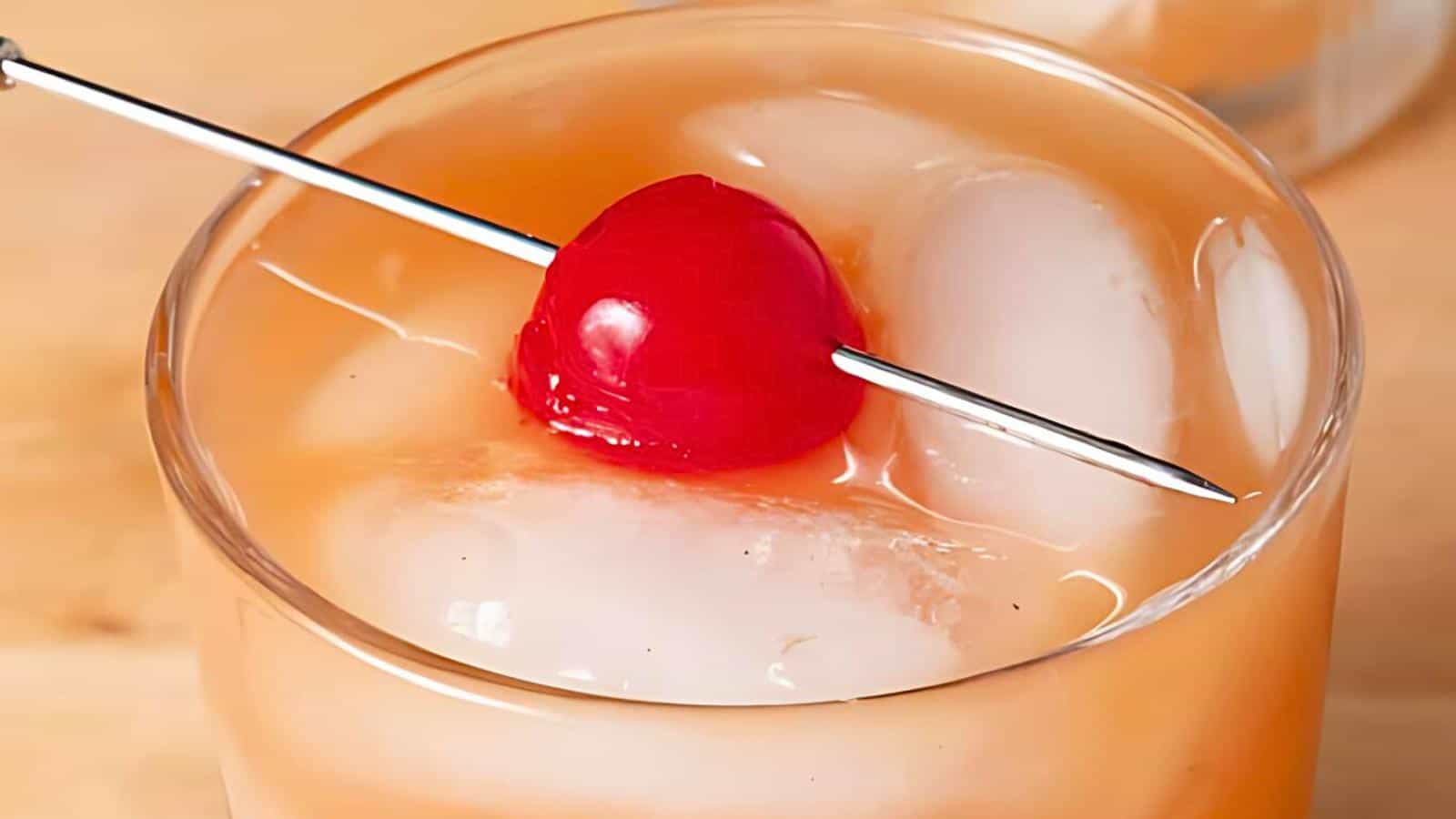A cocktail with ice, a cherry on a skewer on top of the glass, and an orange-tinted liquid.
