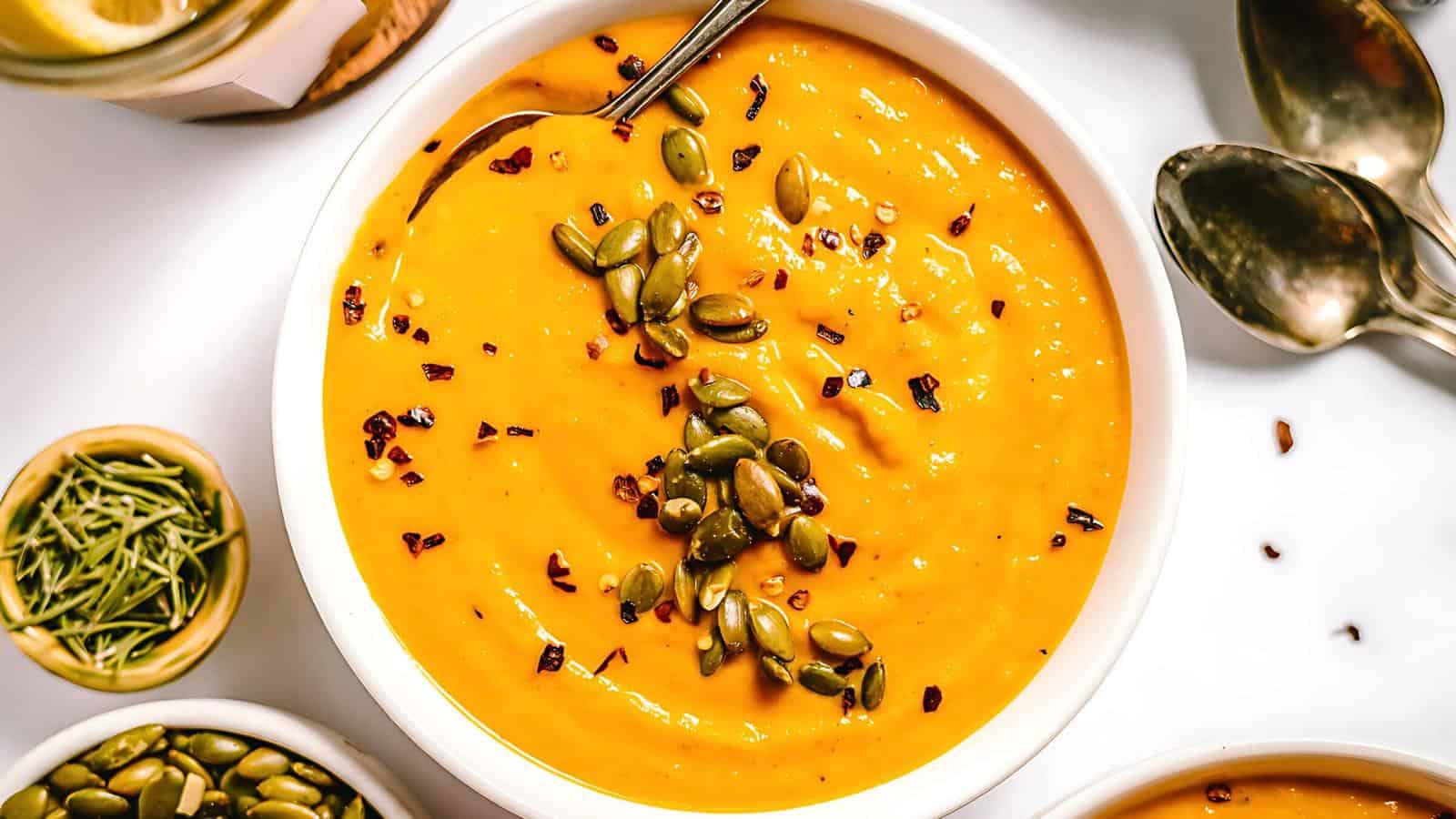 A bowl of soup topped with pumpkin seeds and red chili flakes, surrounded by spoons and small bowls containing various ingredients.