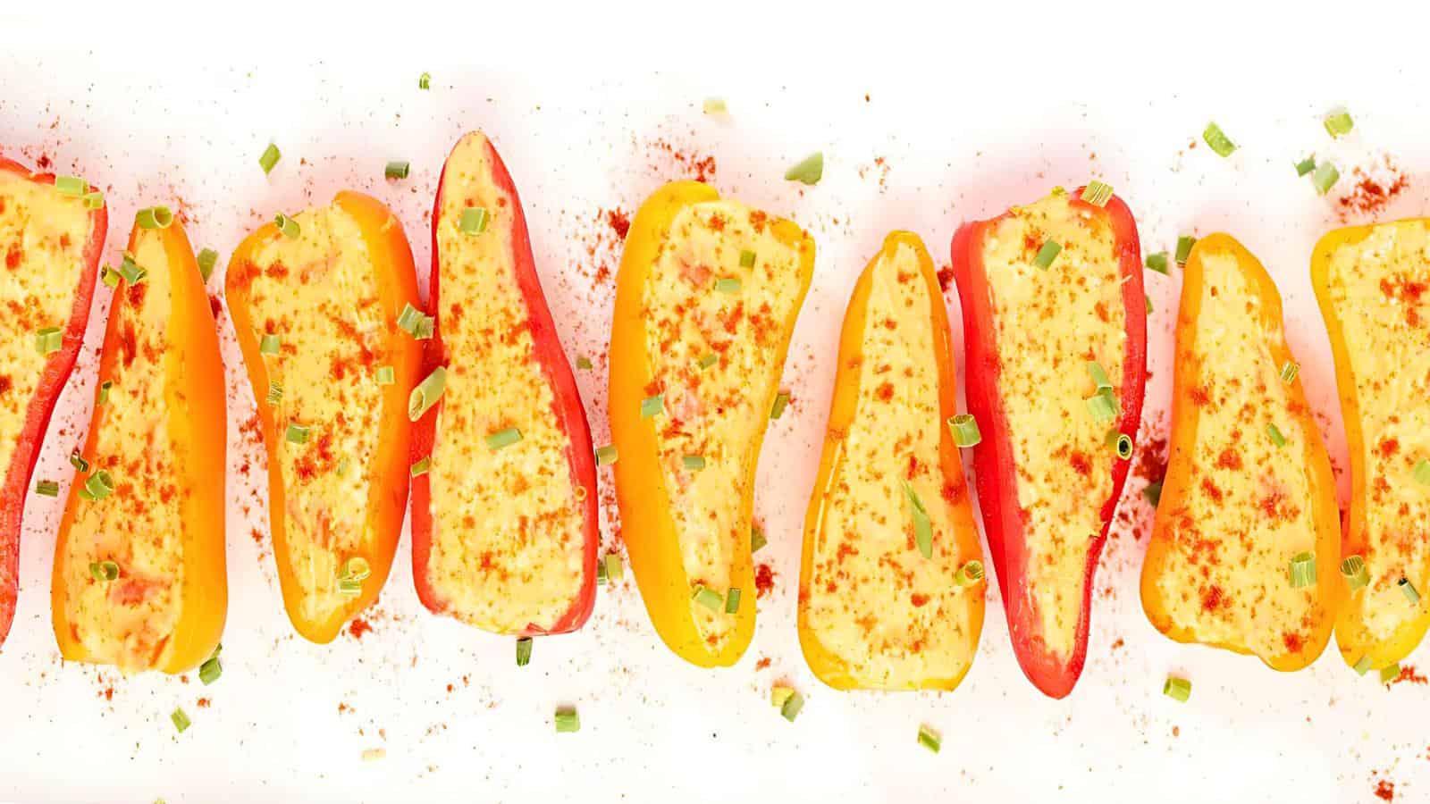Halved mini bell peppers filled with a creamy mixture, sprinkled with paprika and chopped chives, arranged in a row on a white surface.