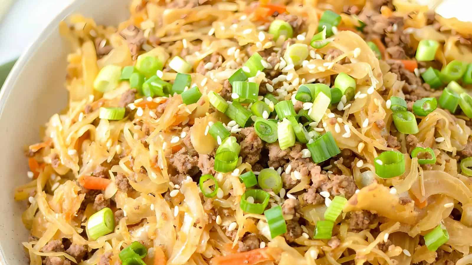 A close-up of a savory dish featuring cooked ground meat mixed with shredded cabbage, sliced onions, and julienned carrots. The dish is garnished with chopped green onions and sprinkled with sesame seeds.