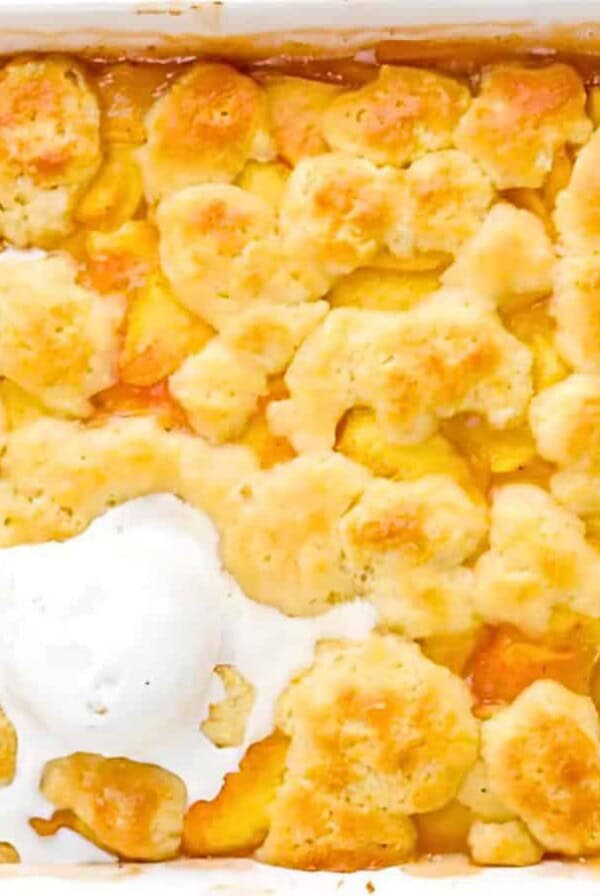 A white baking dish containing baked peach cobbler, topped with three scoops of vanilla ice cream, with a portion of the cobbler served out.