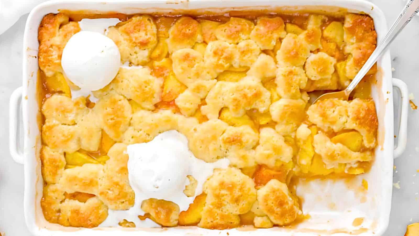 A peach cobbler is shown in a white rectangular baking dish. The cobbler has a golden-brown, unevenly spread crust with patches of fluffy dough. Two scoops of vanilla ice cream are partially melting on top. A spoon rests on the right side of the dish.