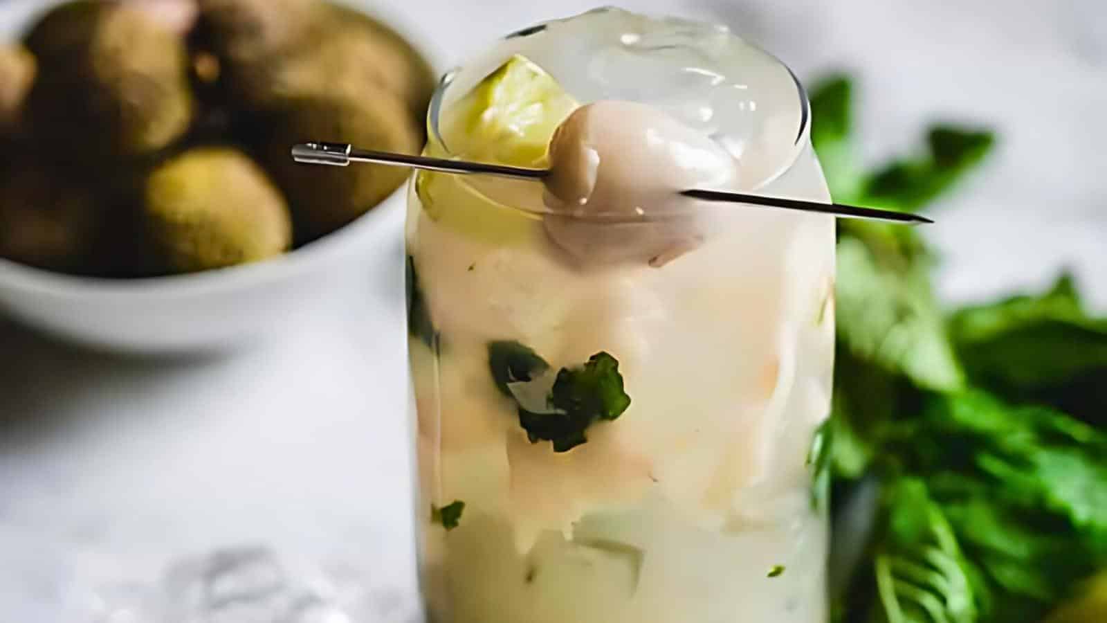 A cocktail garnished with a peeled lychee on a skewer, with a bowl of whole lychees and mint leaves in the background.