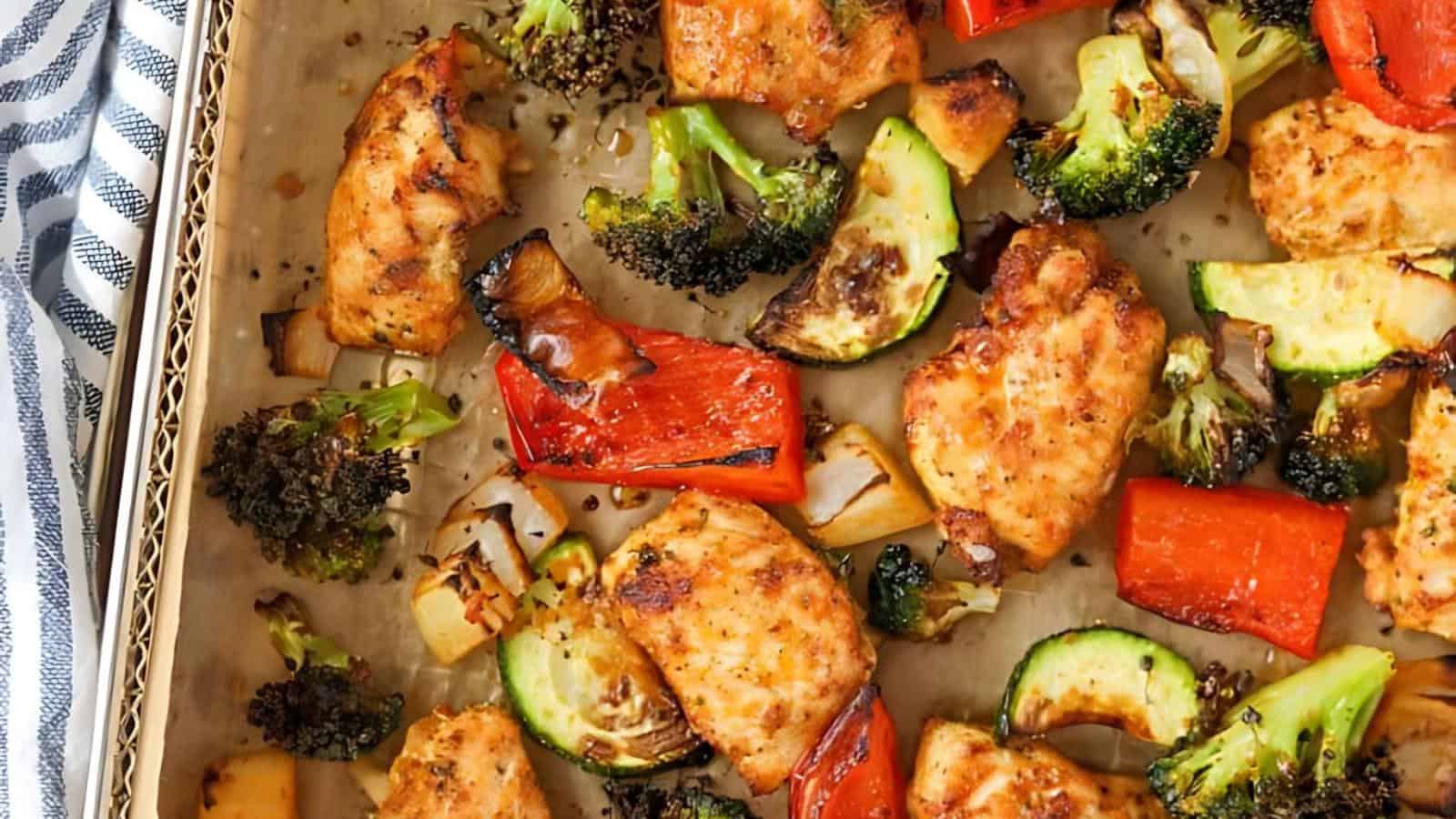 A baking tray filled with cooked chicken pieces, red bell peppers, zucchini slices, broccoli florets, and onions. The food is seasoned and roasted, displaying a golden-brown color, and is positioned on a parchment-lined tray with a striped kitchen towel nearby.