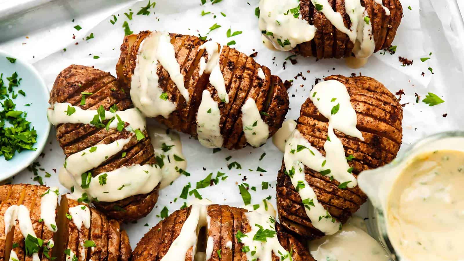 Baked Hasselback potatoes topped with a creamy white sauce and garnished with chopped parsley on a white baking sheet.