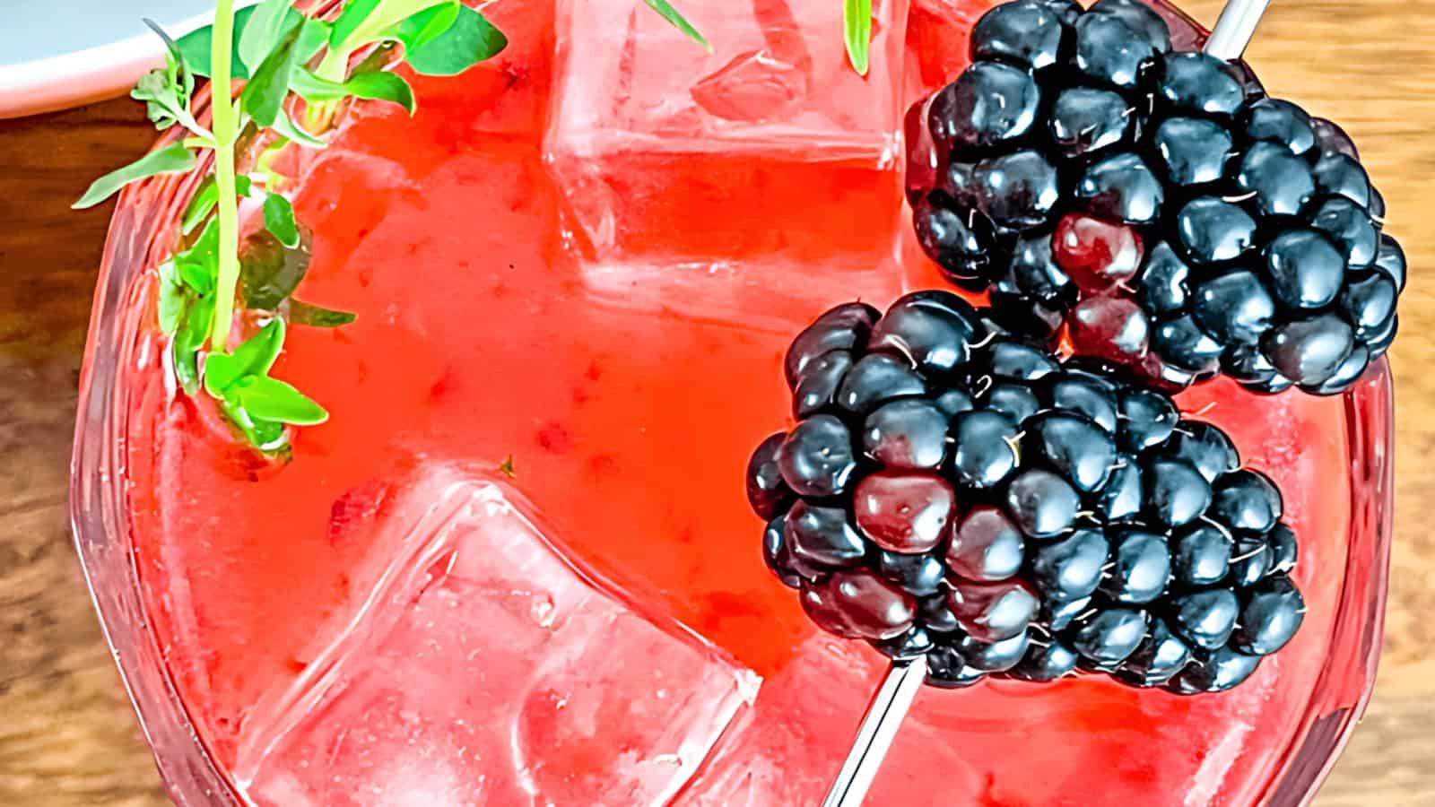 A red cocktail with ice cubes, garnished with two blackberries and a sprig of fresh green herb.