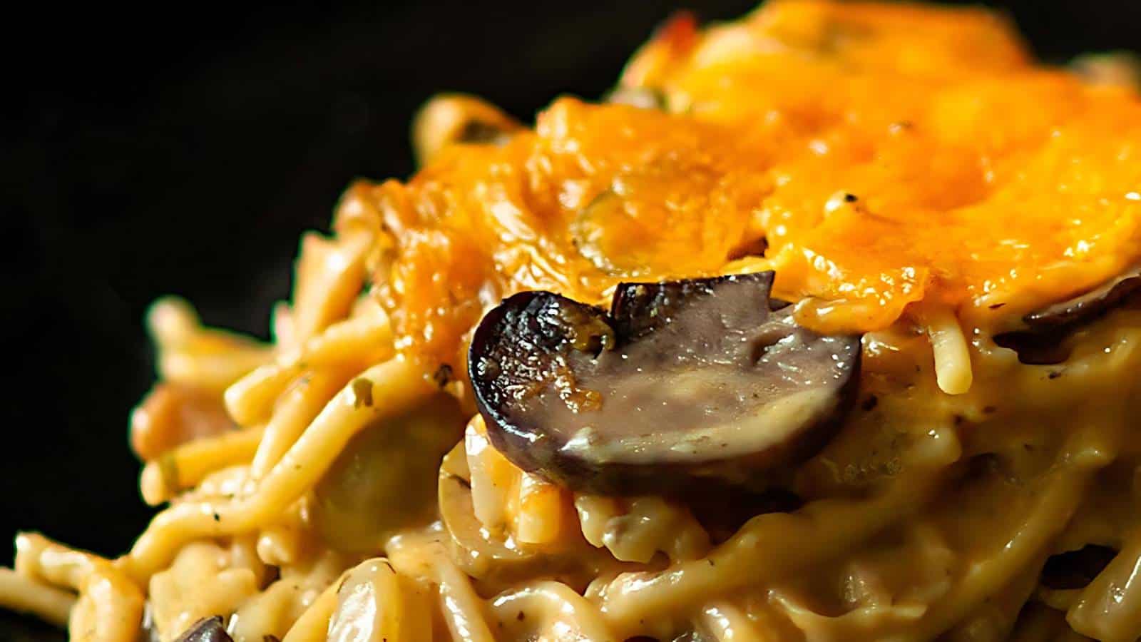 Close-up of a baked pasta dish featuring creamy noodles, sliced mushrooms, and melted cheese with a golden-brown crust. The dish is set against a dark background, highlighting the texture and rich colors of the baked ingredients.