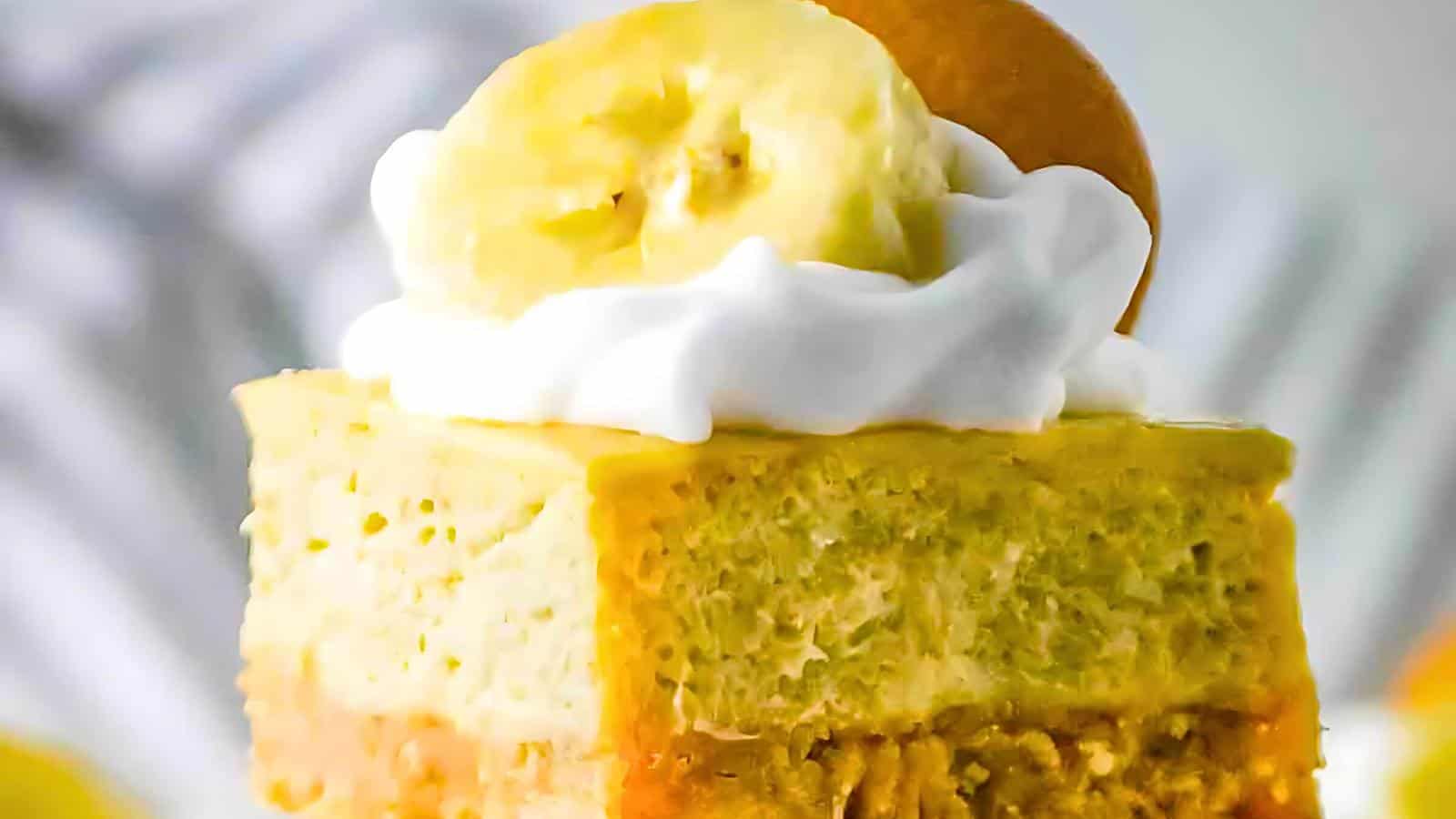 Close-up of a slice of banana pudding cheesecake topped with whipped cream and a banana slice. The creamy, layered dessert has a yellow hue, with distinct layers of pudding, cheesecake, and a crumbly base, garnished with a vanilla wafer. Background is blurred.