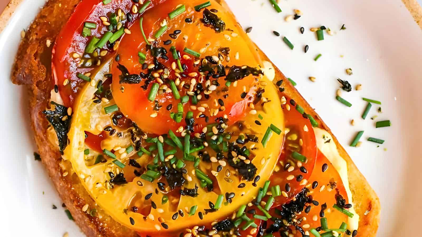 A slice of toast topped with yellow and red tomato slices, chopped chives, sesame seeds, and seasonings on a white plate.