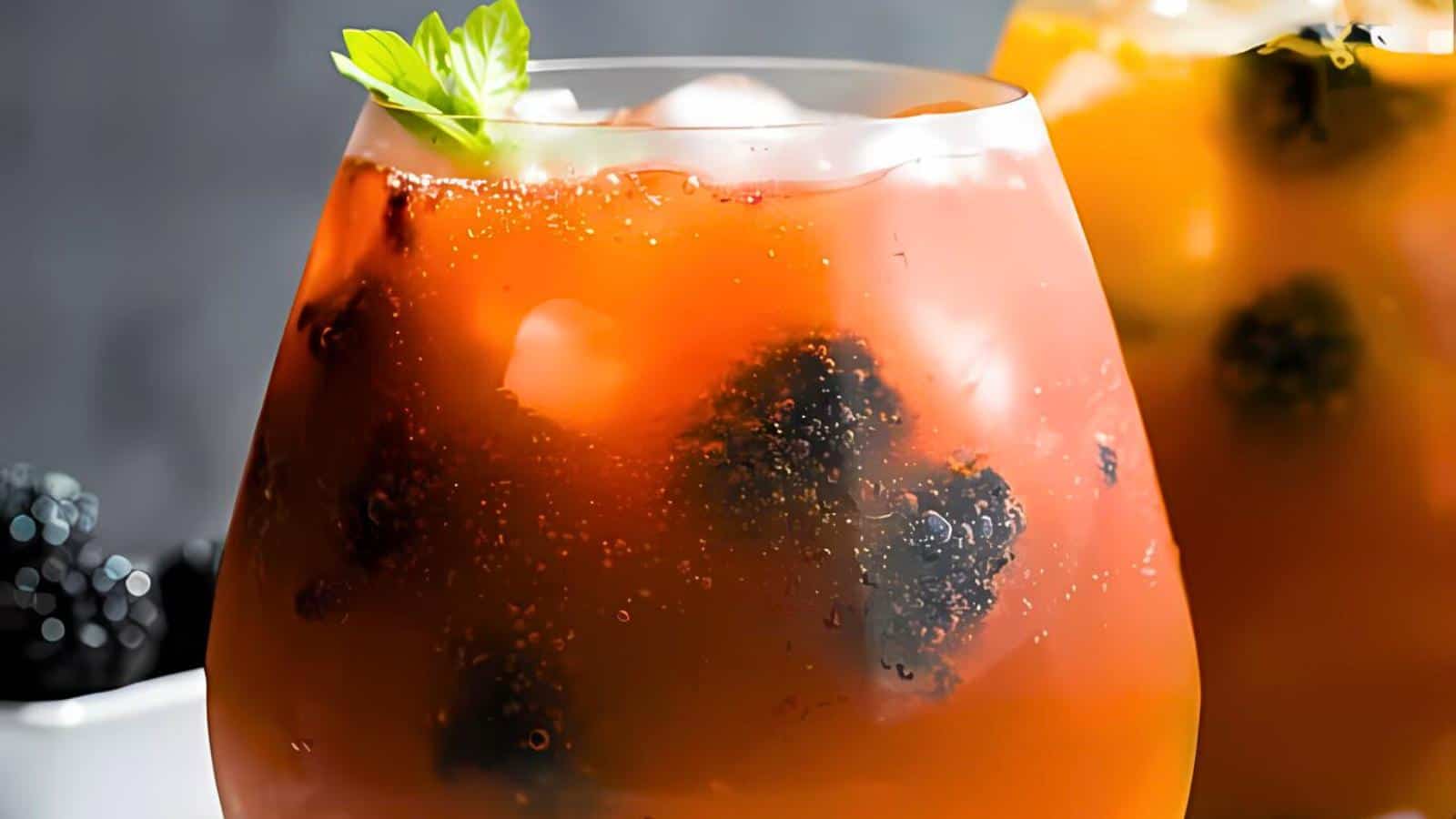 A glass filled with an orange-colored beverage containing ice cubes, blackberries, and garnished with a green leaf. Another similar glass is partially visible in the background.