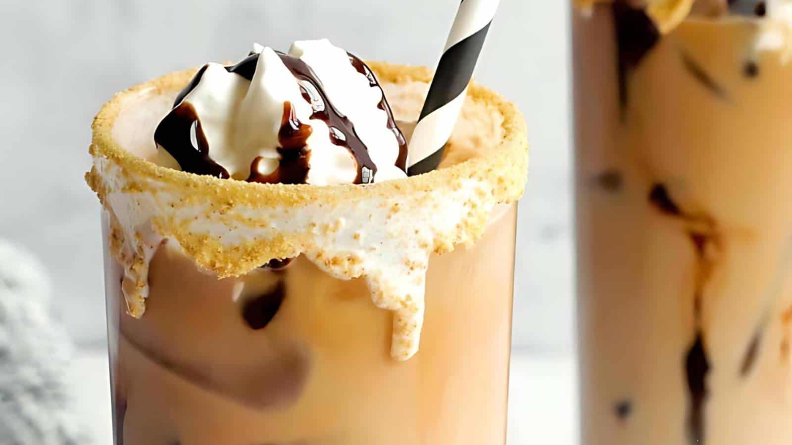 A glass of creamy iced coffee topped with whipped cream and chocolate syrup, featuring a graham cracker rim and a striped straw.