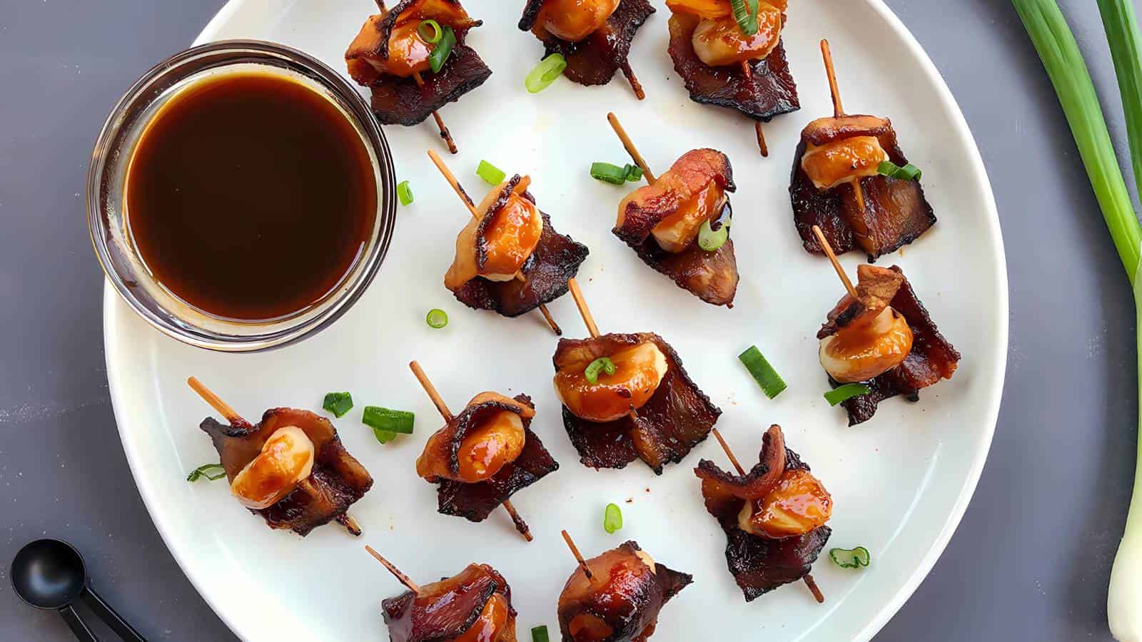 A white plate with bite-sized bacon-wrapped scallops on toothpicks, garnished with chopped green onions, and a small bowl of dark dipping sauce on the side.