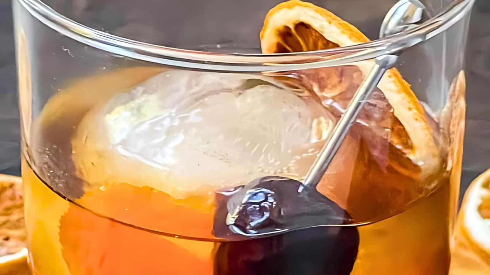 A cocktail in a glass with a large ice sphere, garnished with a dark cherry on a metal skewer and a dried orange slice.