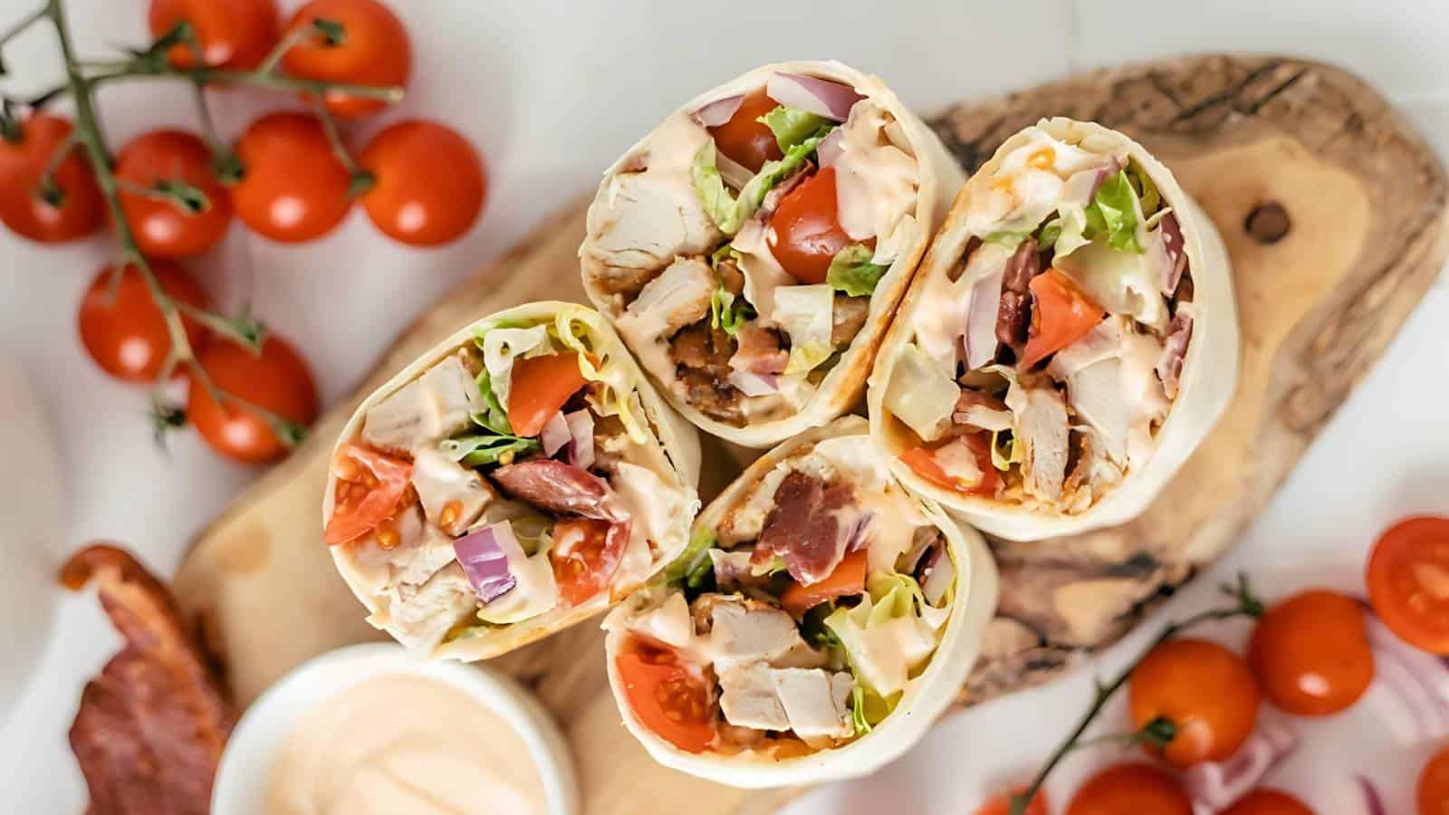 Four chicken wraps filled with vegetables and sauce are arranged on a wooden board, accompanied by cherry tomatoes and a small dish of dipping sauce.