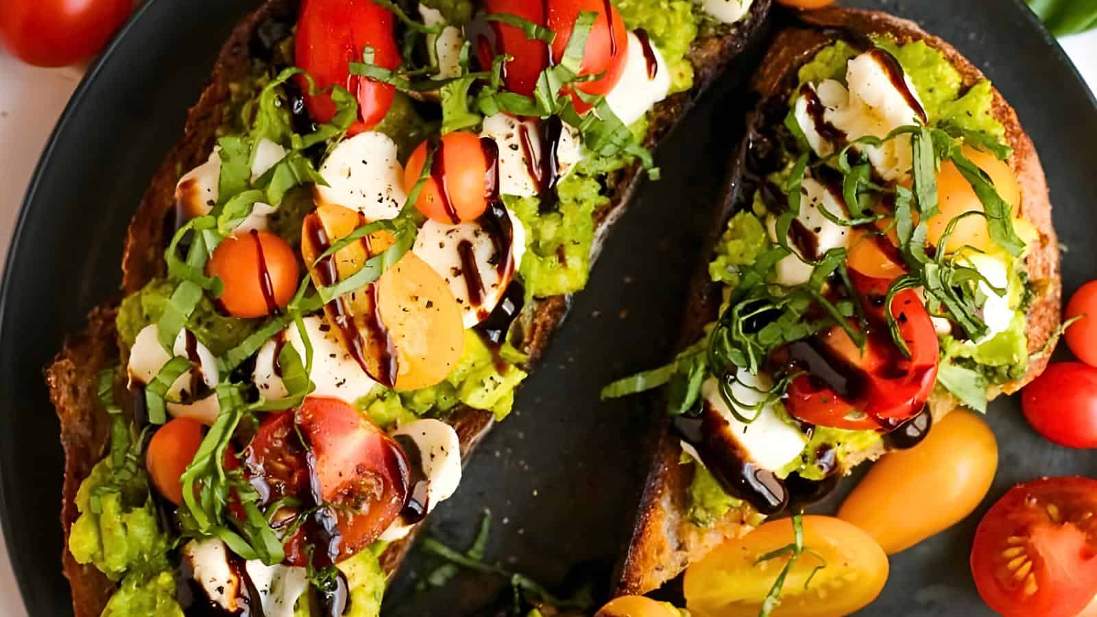 Avocado toast topped with cherry tomatoes, mozzarella pieces, chopped basil, and drizzled with balsamic glaze on a black plate.
