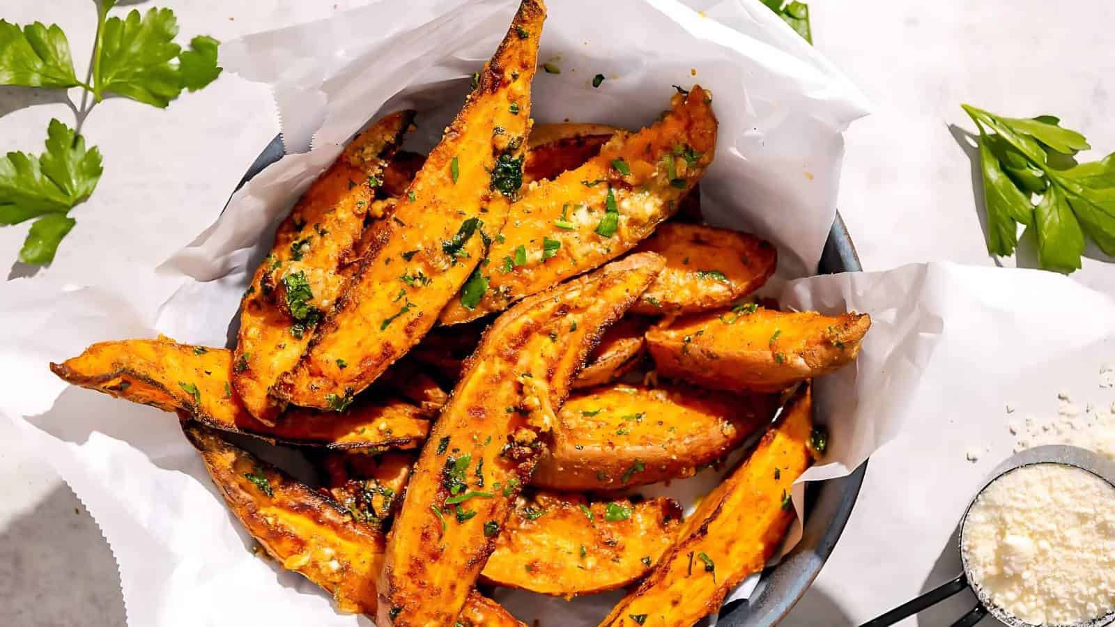 Seasoned and baked sweet potato wedges garnished with fresh parsley, placed on white parchment paper. Parsley leaves and grated cheese are scattered around.