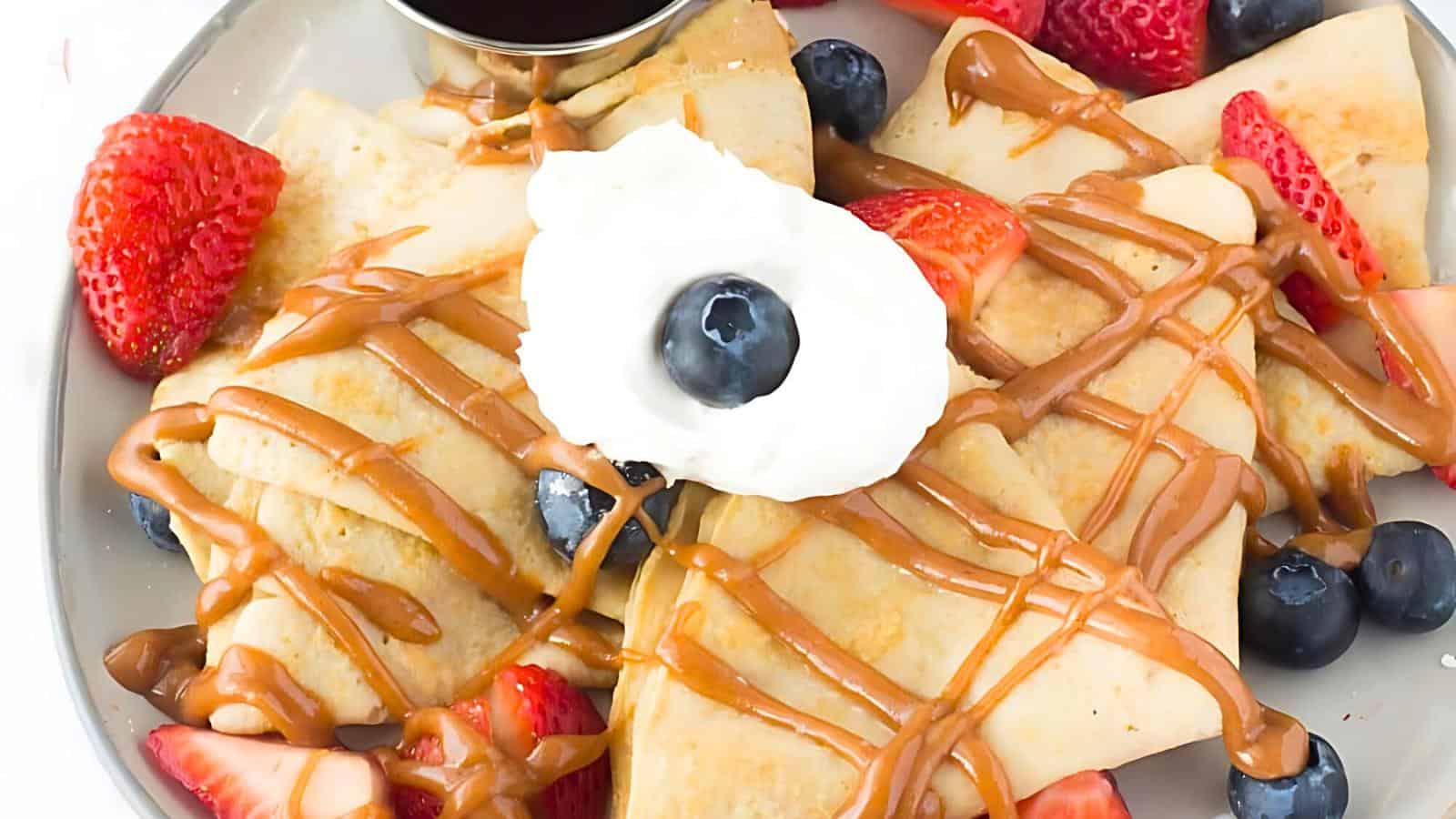 A plate of crepes drizzled with caramel sauce, topped with whipped cream, blueberries, and cut strawberries with a small cup of sauce on the side.