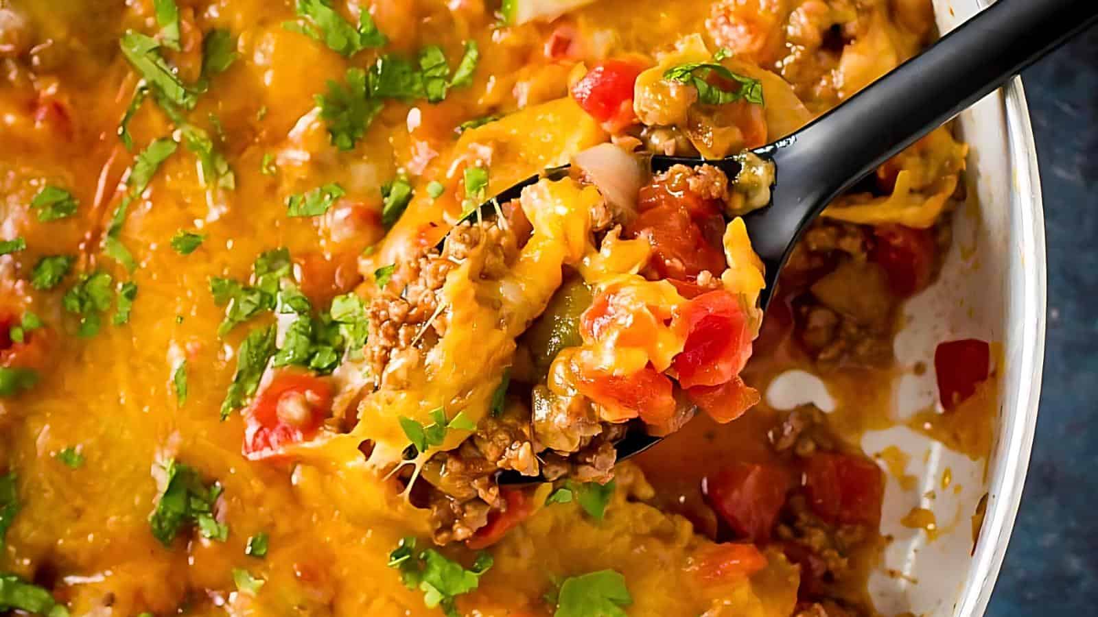 A close-up image of a hearty casserole dish showing a spoonful being lifted. The dish is topped with melted cheese and garnished with fresh chopped herbs, revealing a mix of ground meat, diced tomatoes, and other ingredients.