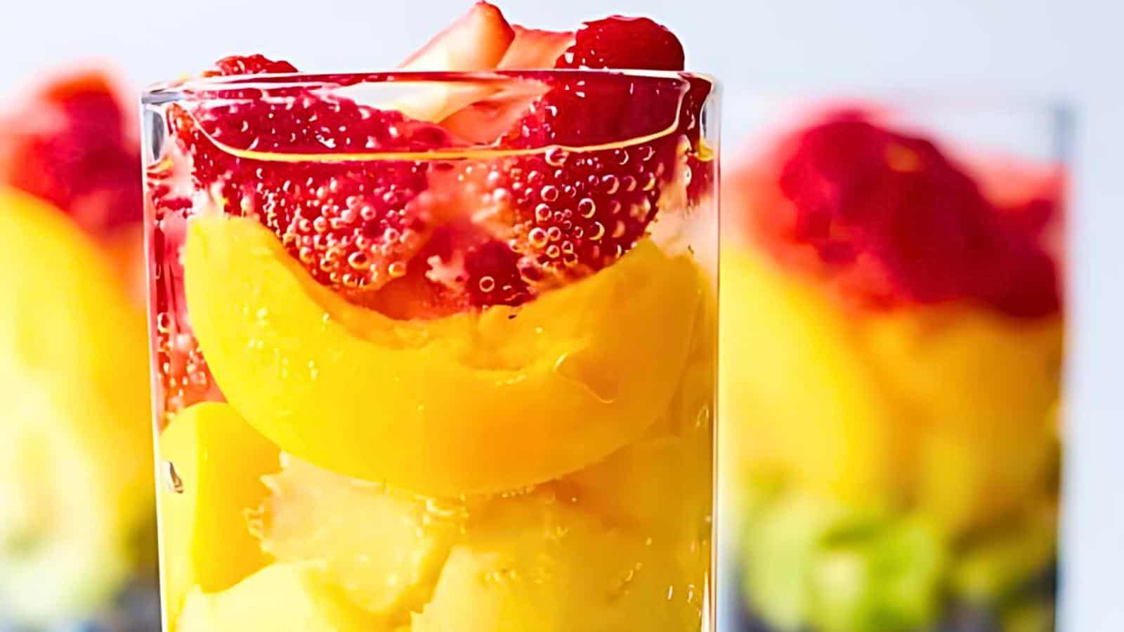 A glass filled with layered fresh fruits: strawberries, peach slices, and kiwi chunks. The background shows similar glasses out of focus.