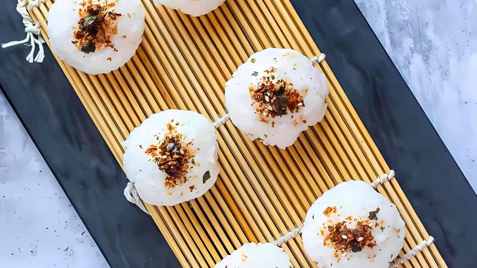 Six rice balls topped with seasoning are arranged diagonally on a bamboo mat and placed on a black rectangular plate.