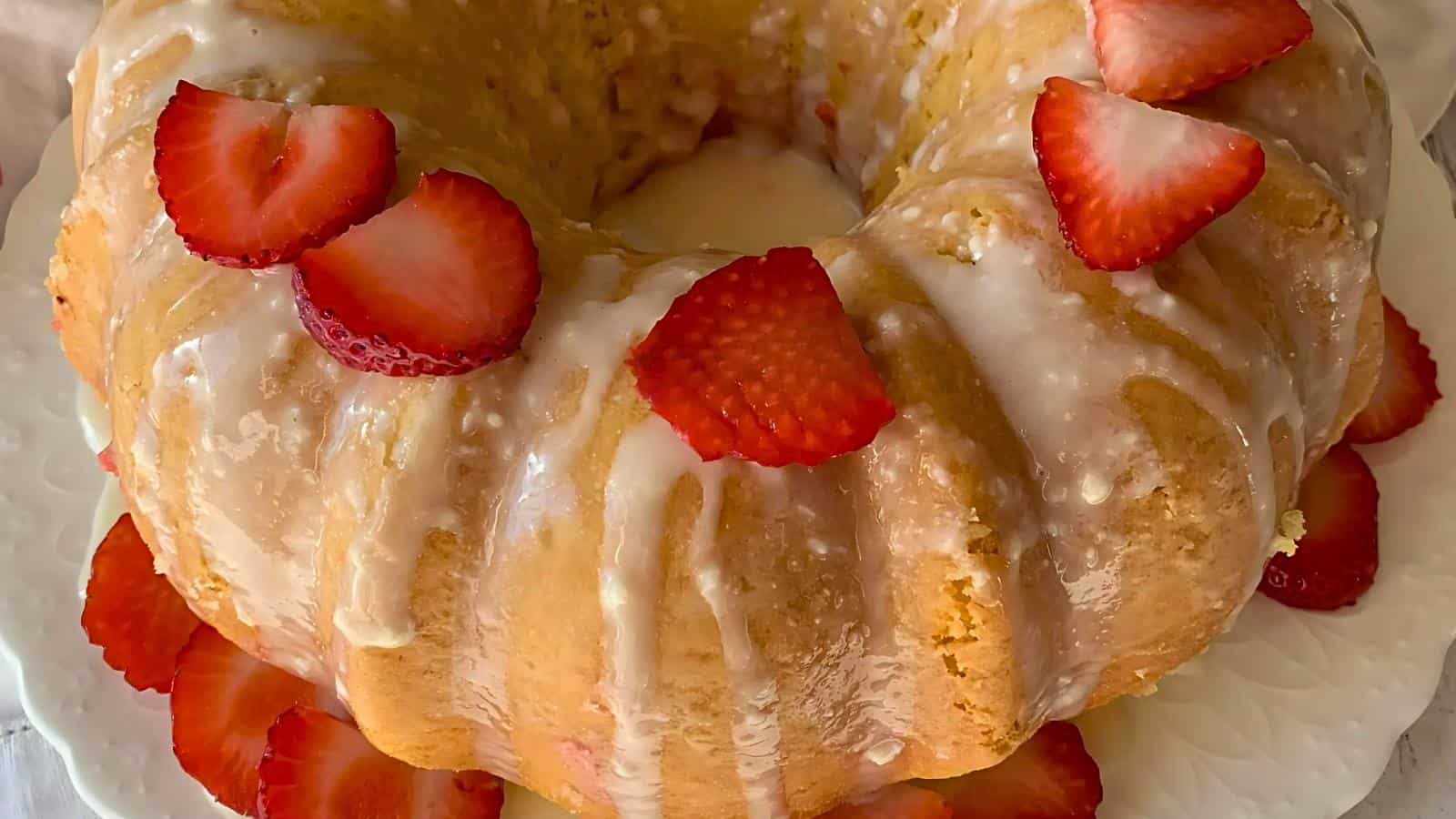A glazed poke cake topped with fresh strawberry slices on a white plate, with additional strawberry slices arranged around the base of the cake.