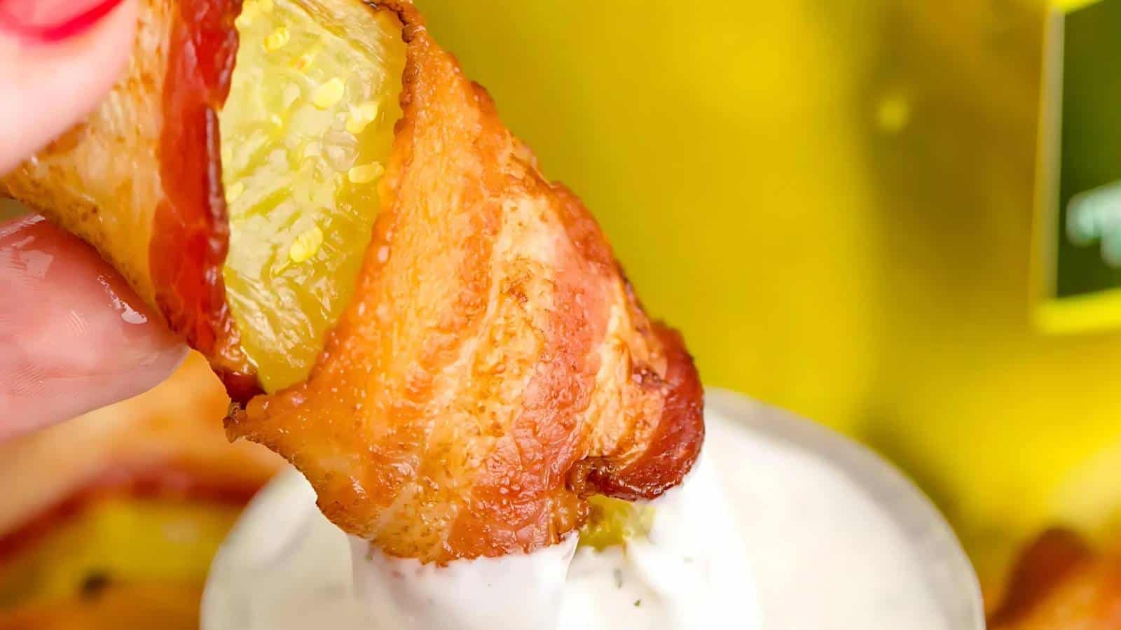 A bacon-wrapped pickle being dipped into a white sauce.