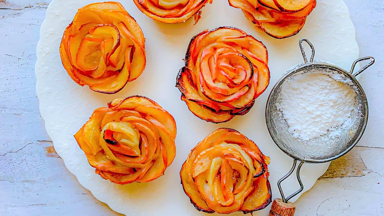 Plate with apple rose pastries and a sifter filled with powdered sugar on a light wooden surface.