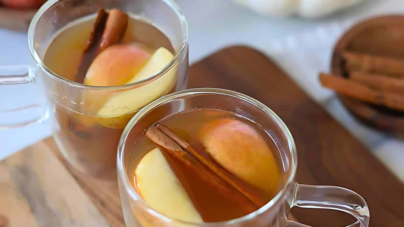 Two clear glass mugs filled with an apple cider, each garnished with apple slices and cinnamon sticks, on a wooden surface.