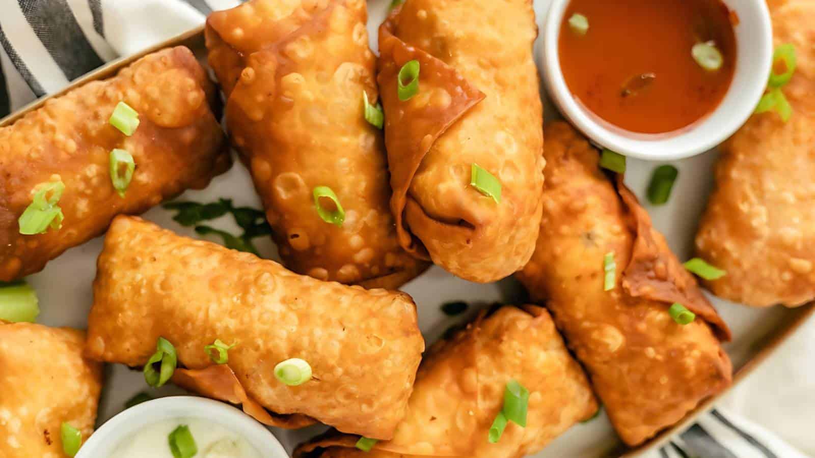A plate of golden-brown egg rolls garnished with chopped green onions, accompanied by two small bowls of dipping sauces.