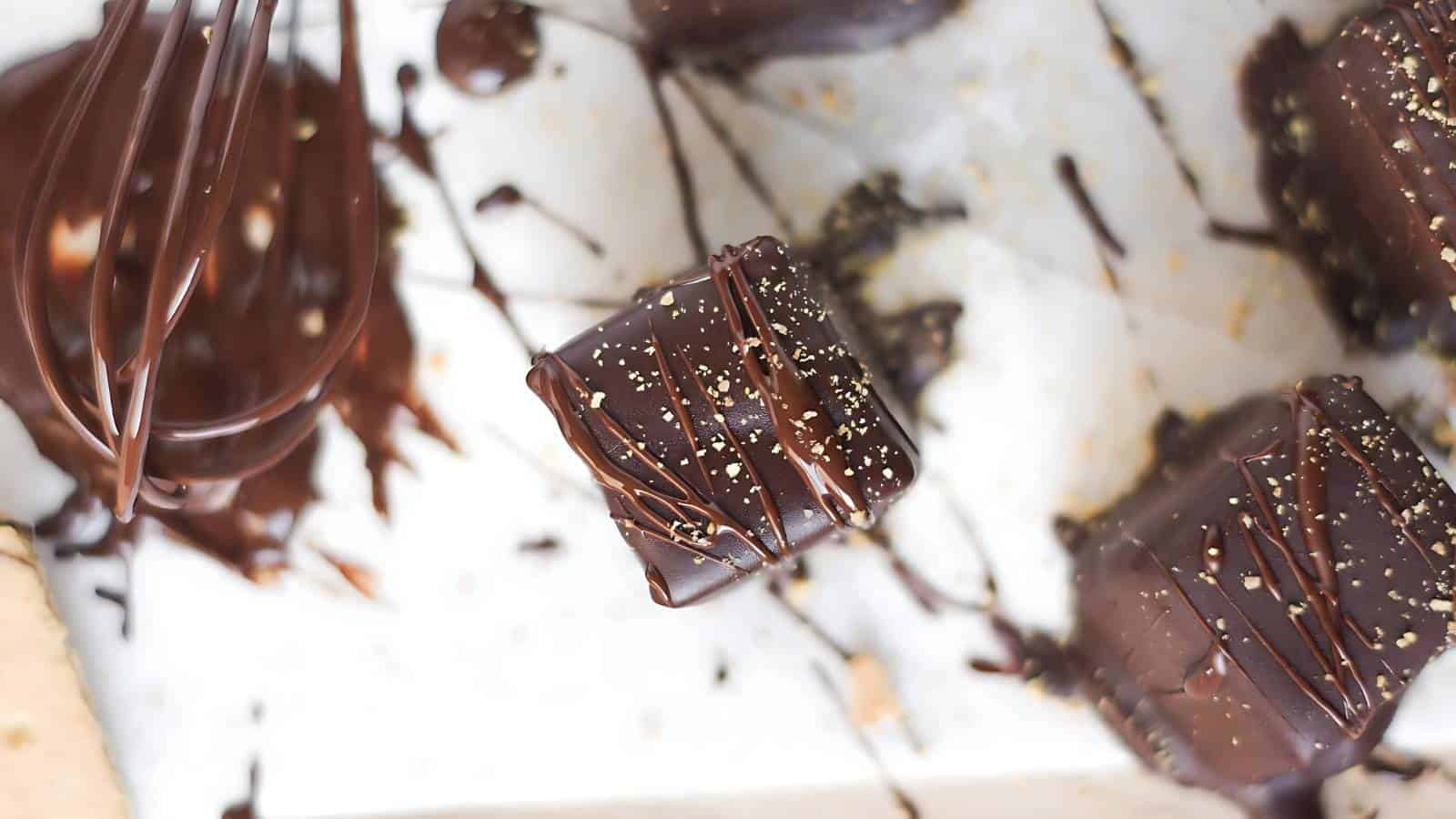 Chocolate-covered square treats with drizzled chocolate and sprinkles, a whisk coated with melted chocolate on a light surface.