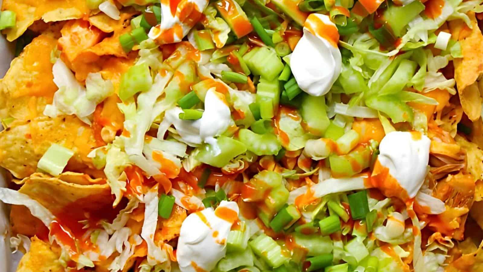 A close-up of a plate of nachos topped with shredded lettuce, chopped green onions, diced celery, small dollops of sour cream, and drizzled with hot sauce. The vibrant colors of the fresh ingredients stand out against the golden color of the nacho chips.