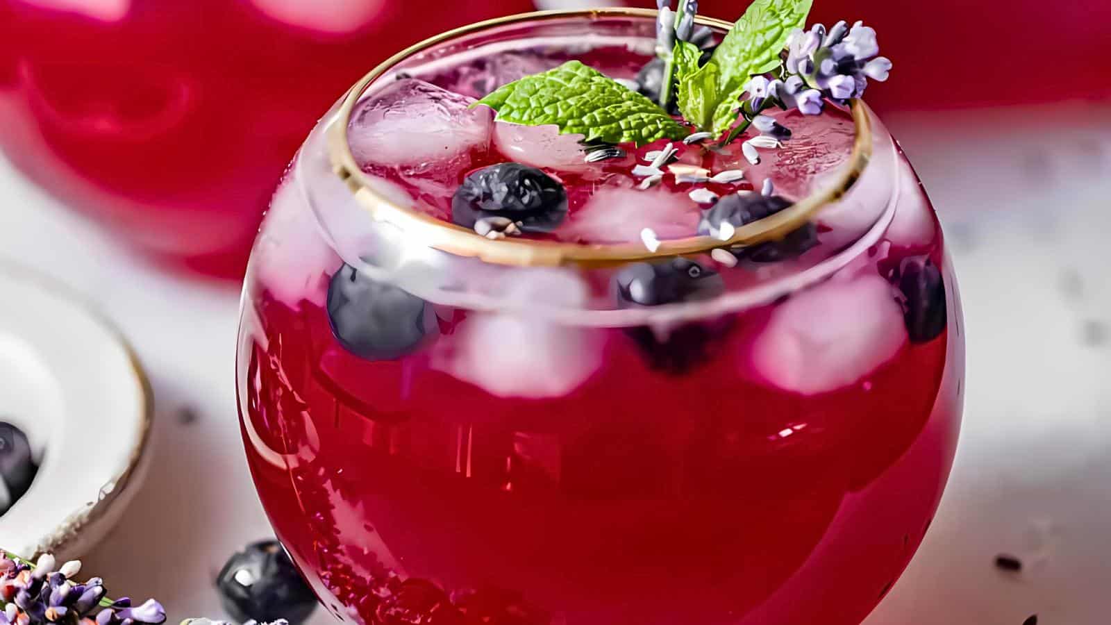 A glass of lemonade with ice cubes, blueberries, mint leaves, and flower petals on top. Another glass partially visible in the background.