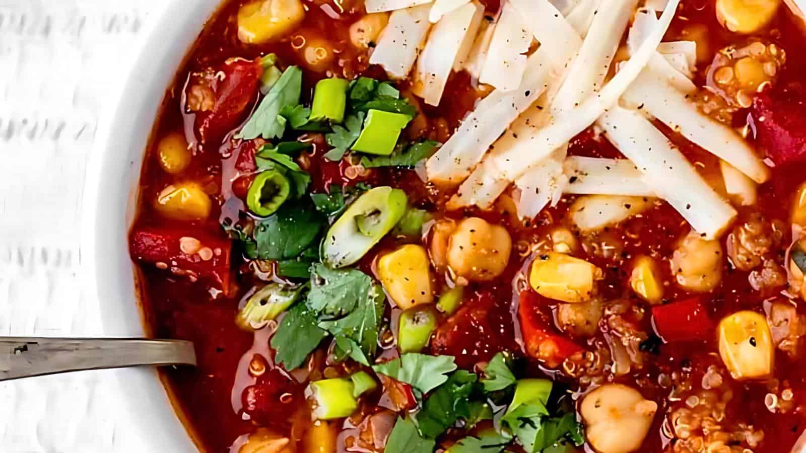 A bowl of soup topped with shredded cheese, chopped green onions, and cilantro. The chili contains corn, tomatoes, beans, and various vegetables.
