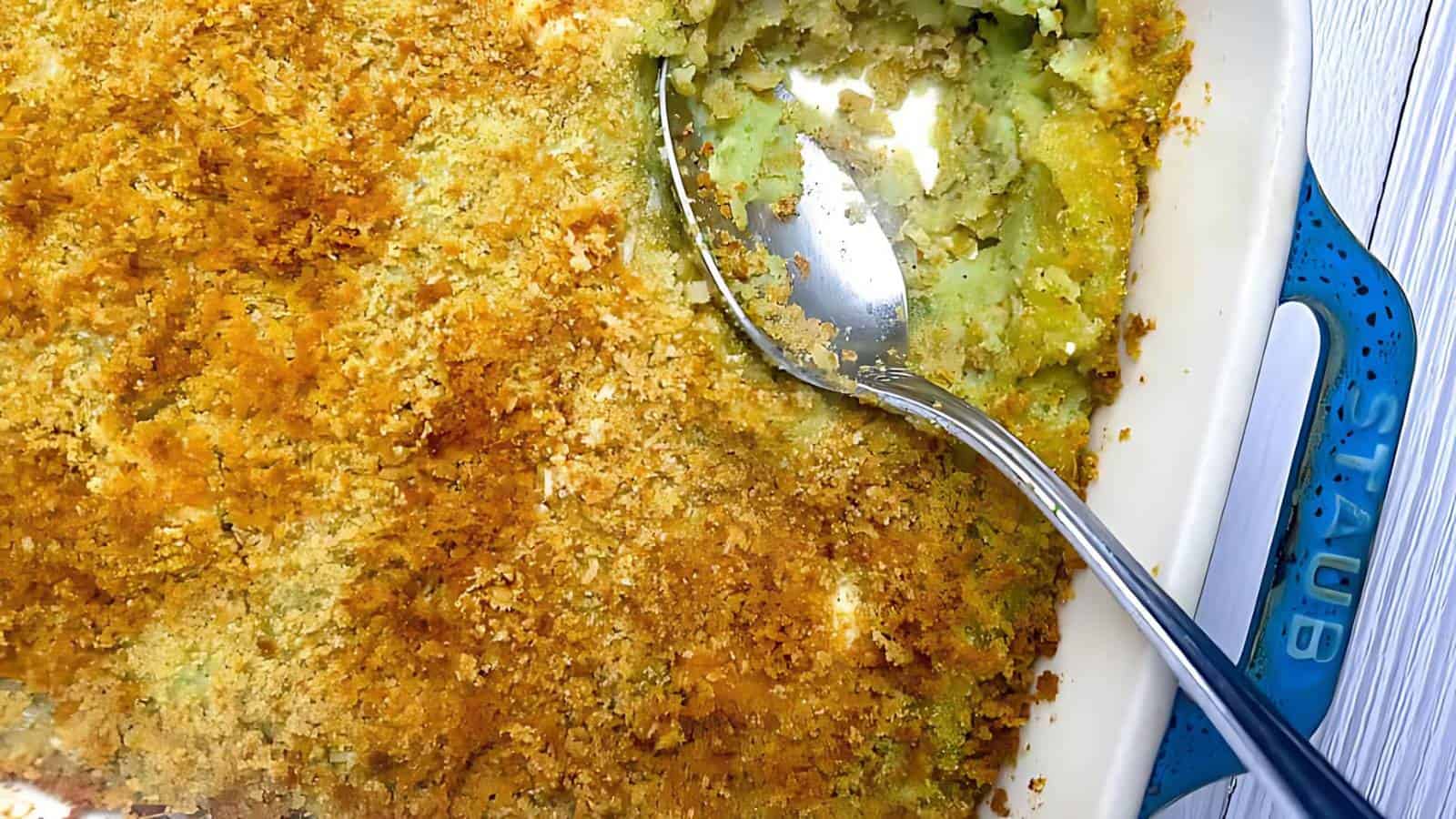 A casserole dish with a bread crumb topping, a serving spoon, and a portion missing, revealing a creamy green filling.