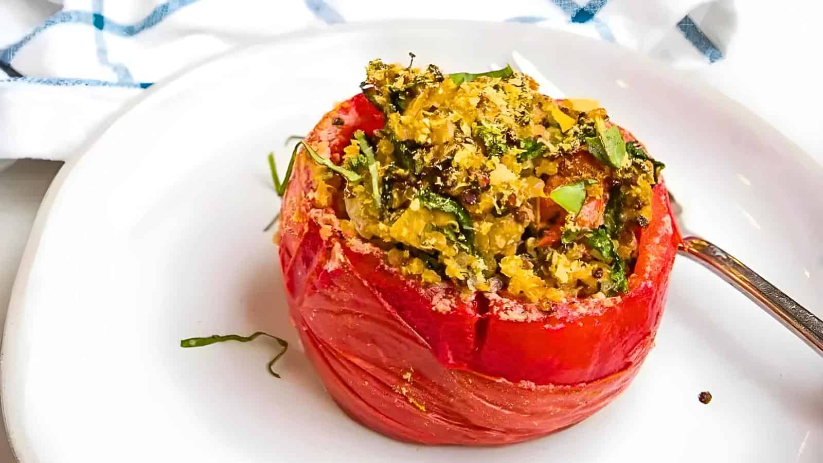 A stuffed tomato filled with quinoa and chopped vegetables sits on a white plate next to a fork.