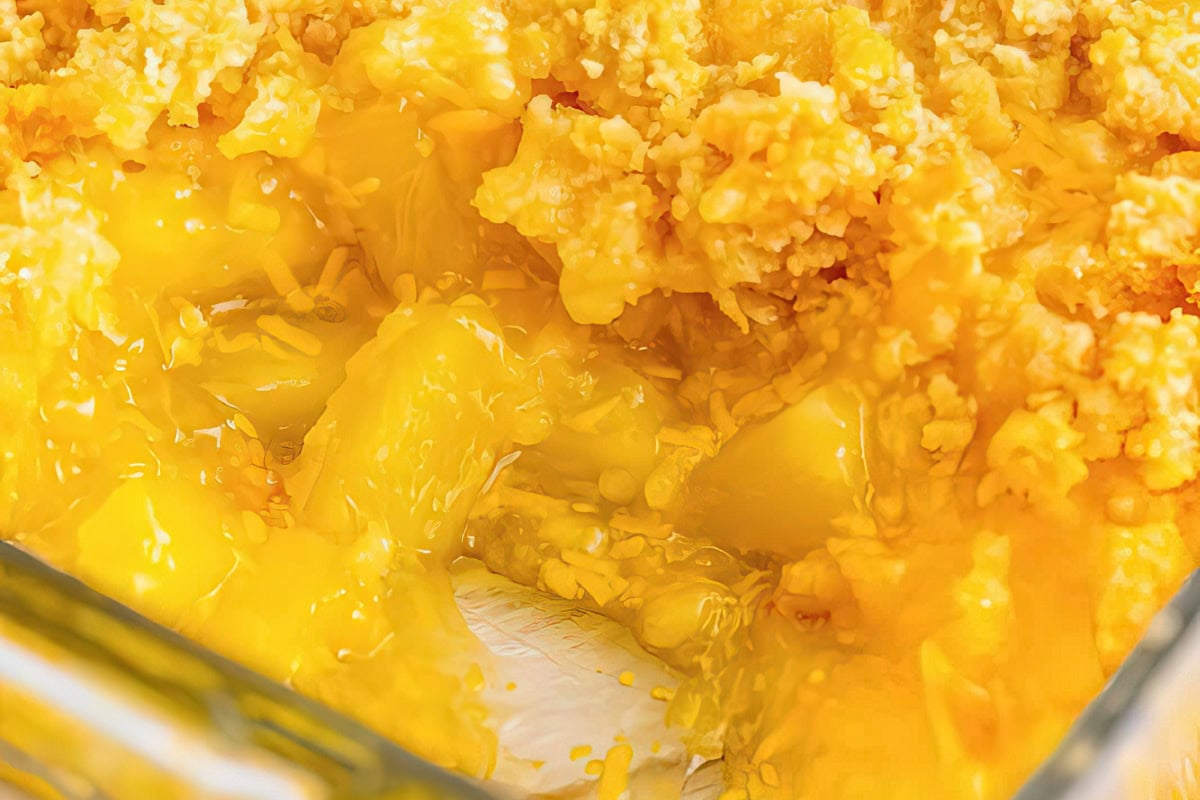A close-up view of a dessert featuring chunks of yellow pineapple coated in a glossy yellow gelatinous substance, topped with a crumbly, golden-brown crust. The dessert is presented in a glass dish.