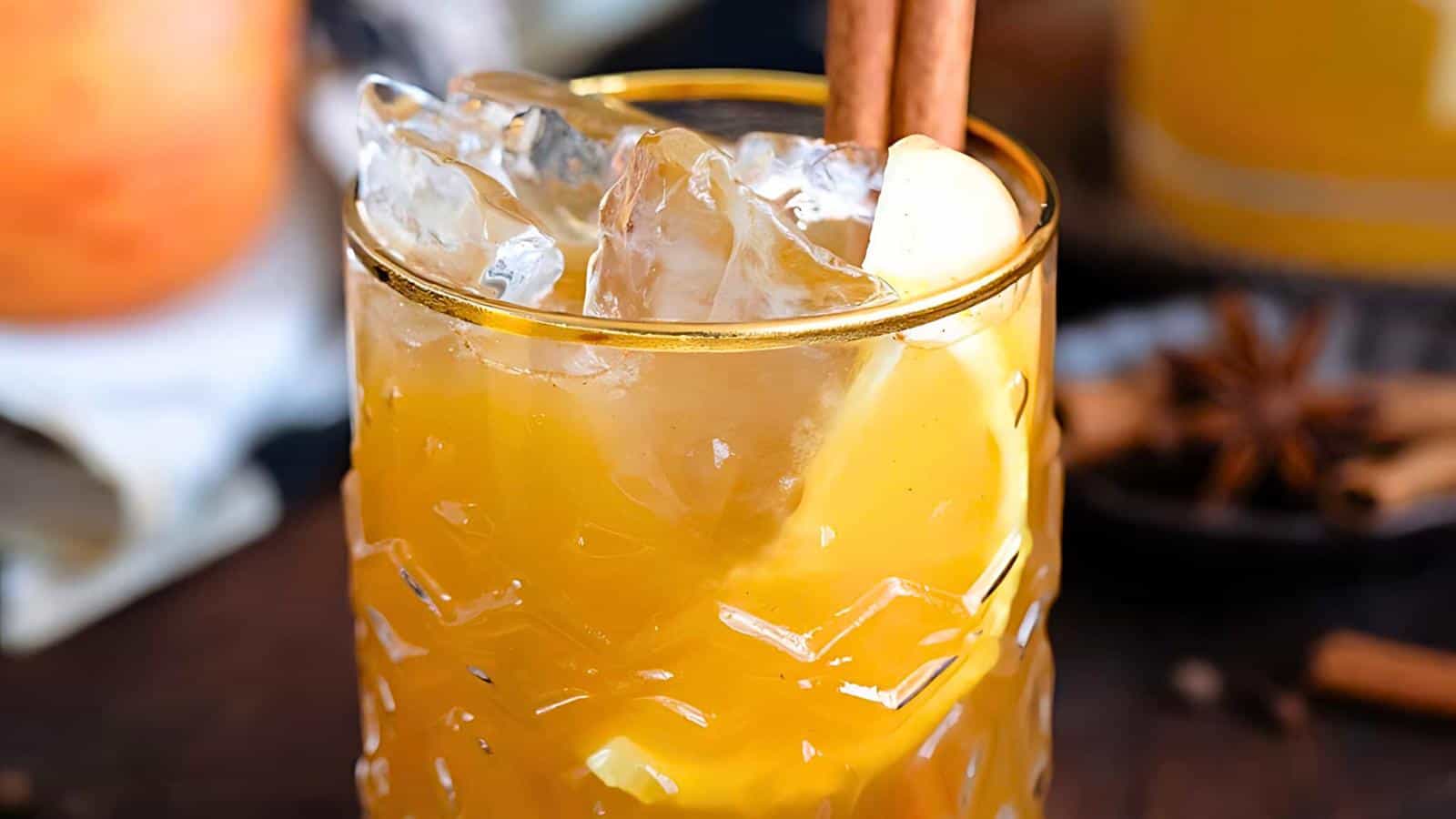 A glass with an orange-yellow beverage, ice cubes, a lemon slice, and a cinnamon stick.
