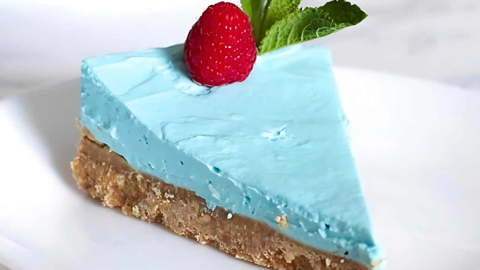A slice of blue cheesecake topped with a raspberry and a sprig of mint sits on a white plate.