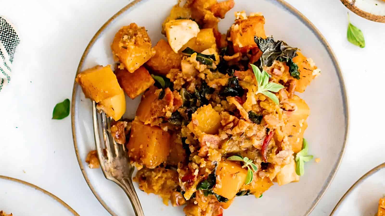 A plate of roasted butternut squash mixed with kale, bacon, and herbs, with a fork placed on the side.