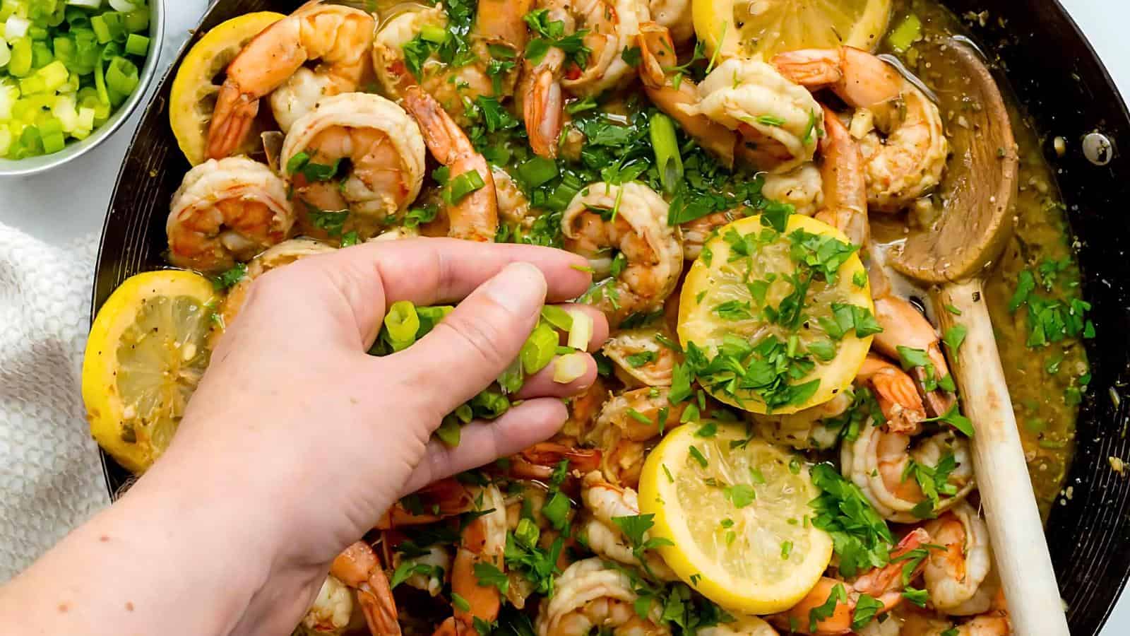 A skillet filled with cooked shrimp, lemon slices, chopped green onions, and fresh herbs. A wooden spoon rests in the skillet.