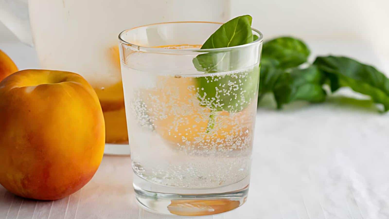 A clear glass of fizzy water garnished with an orange slice and a basil leaf, placed on a table with apricots and fresh basil leaves in the background.
