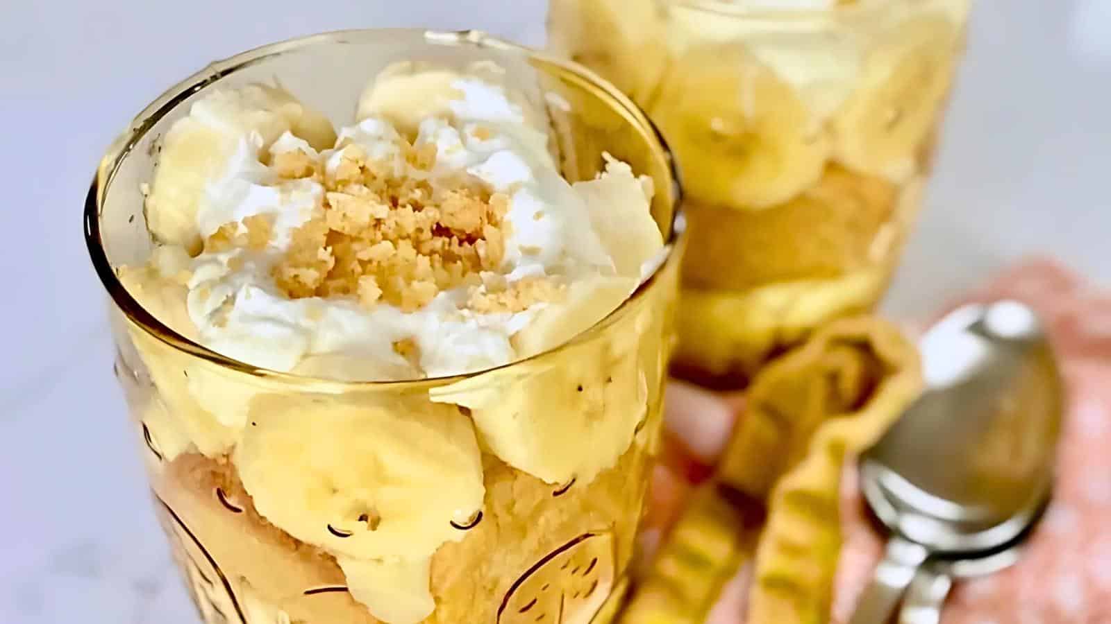 Two glasses filled with creamy banana pudding topped with whipped cream and crushed cookies. The dessert is layered with banana slices and pudding. Metal spoons and a patterned orange napkin are placed beside the glasses on a white surface.