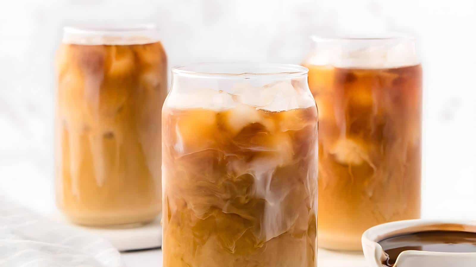 Three glass jars with iced coffee, featuring a blend of coffee and cream with ice cubes. A small bowl of cream in the foreground. Blurred background.