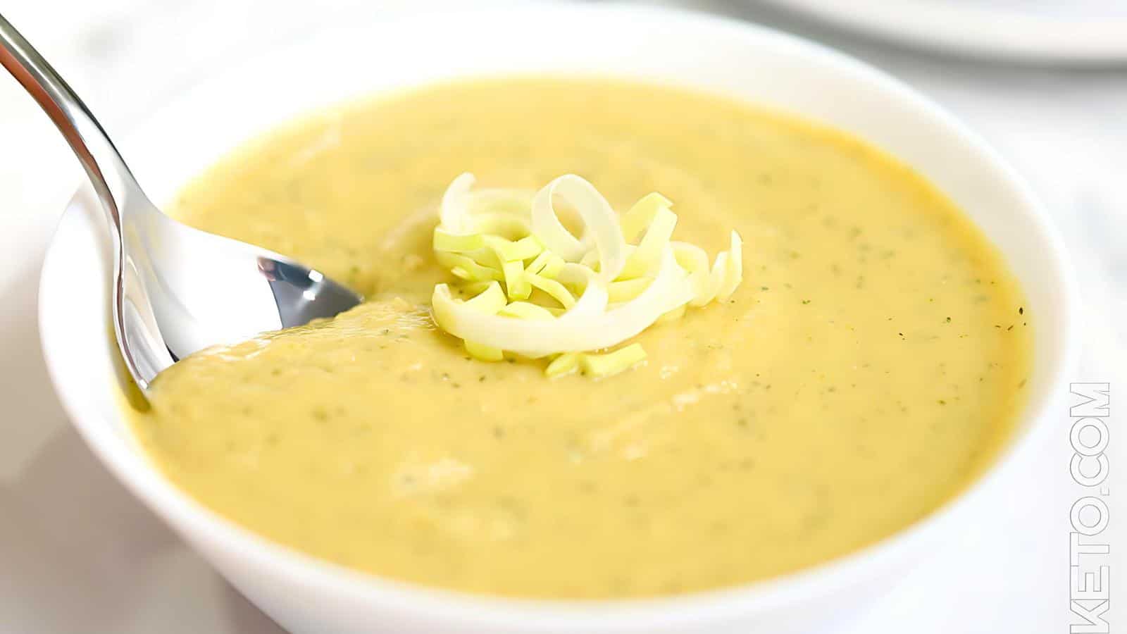 A spoon in a bowl of yellow soup, topped with sliced leeks.