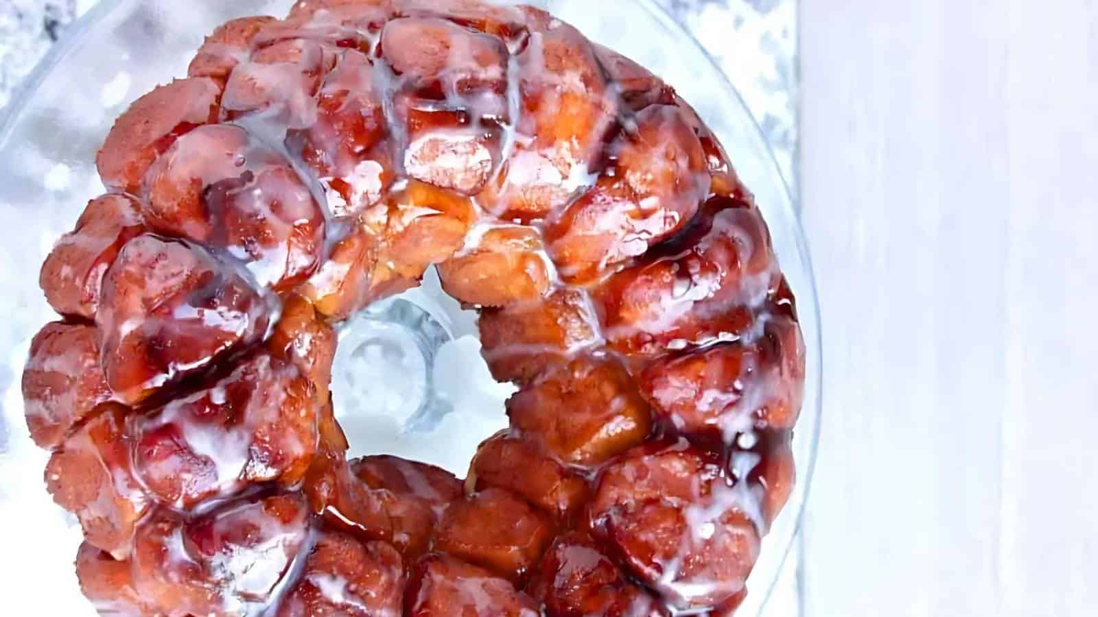 A close-up of a glazed monkey bread on a glass cake stand. The bread is golden brown, with a sticky, shiny glaze dripping down its textured surface, arranged in a circular, pull-apart pattern. The background is softly blurred.