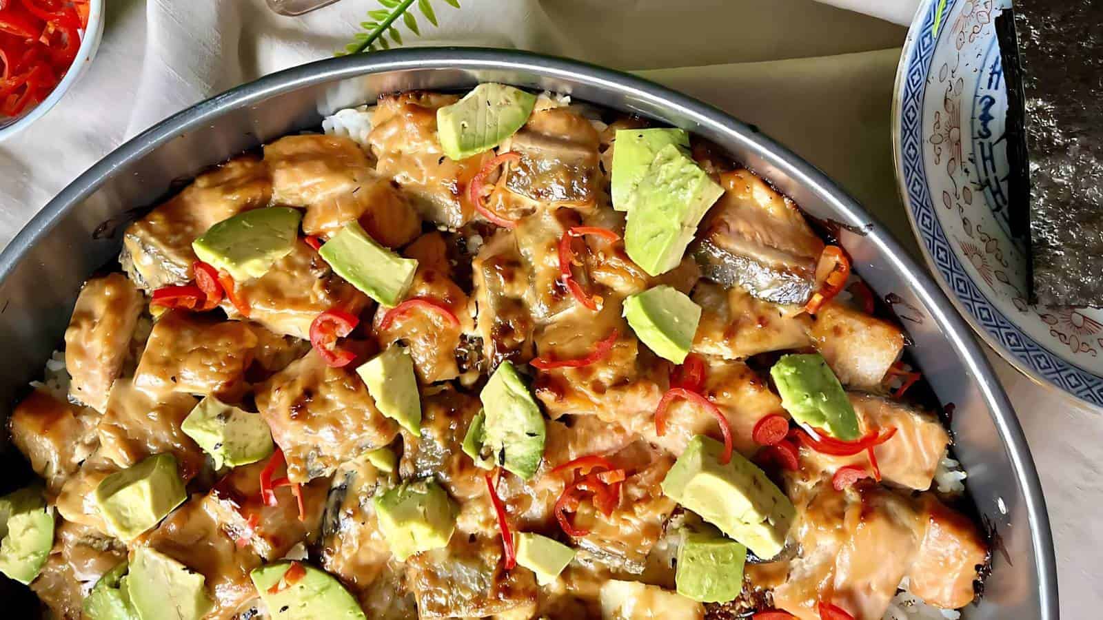 A dish with rice topped with pieces of salmon, avocado, and red pepper slices, garnished with a creamy sauce. China bowls and other ingredients partially visible on the side.
