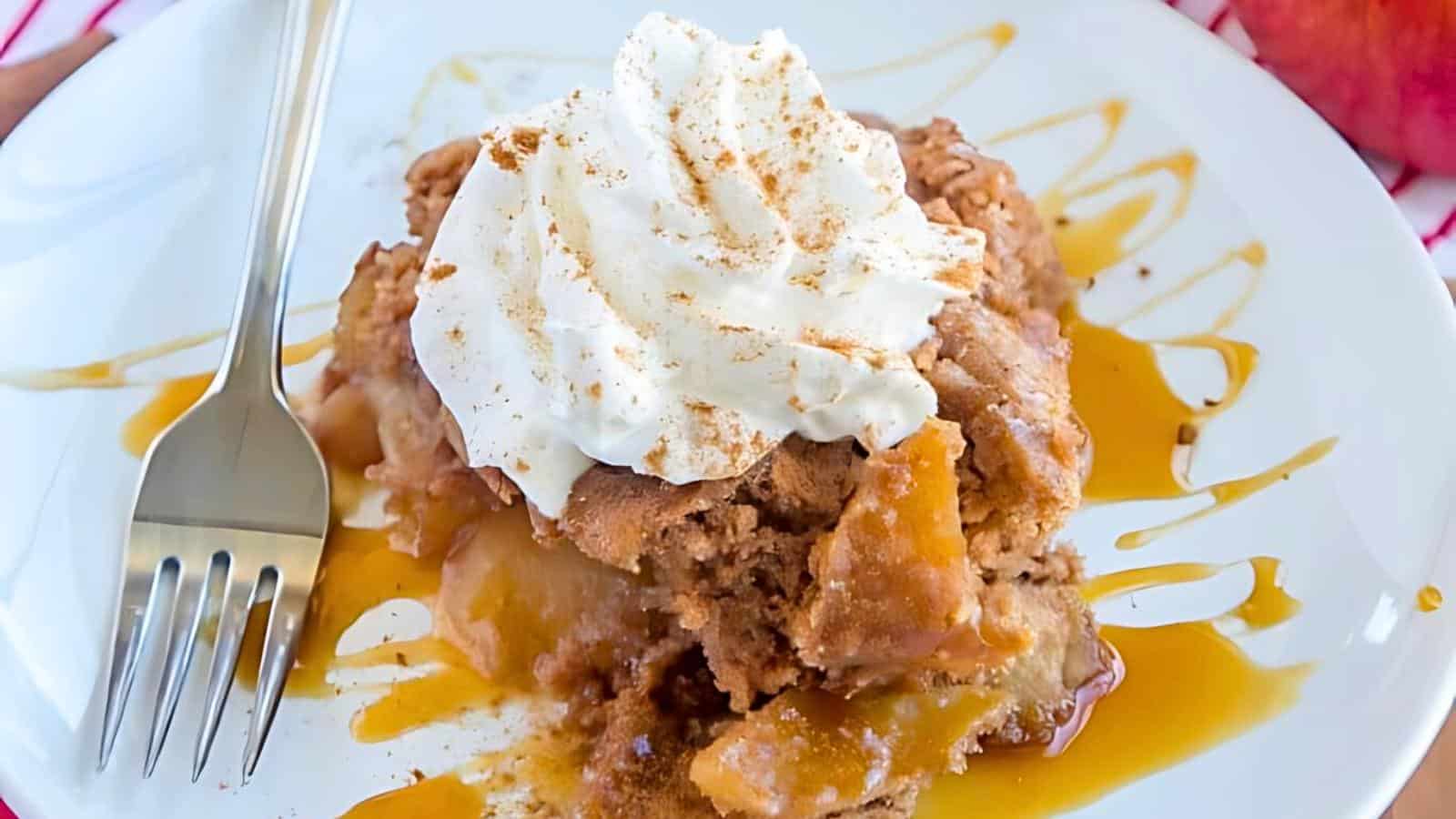 A plate of apple crisp is topped with a dollop of whipped cream and a sprinkle of cinnamon. Caramel drizzle surrounds the dessert, and a fork lies beside it on the white plate.