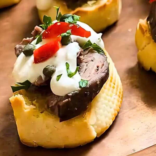 Toasted bread slices topped with meat, cream sauce, chopped red peppers, capers, and parsley on a wooden plate.