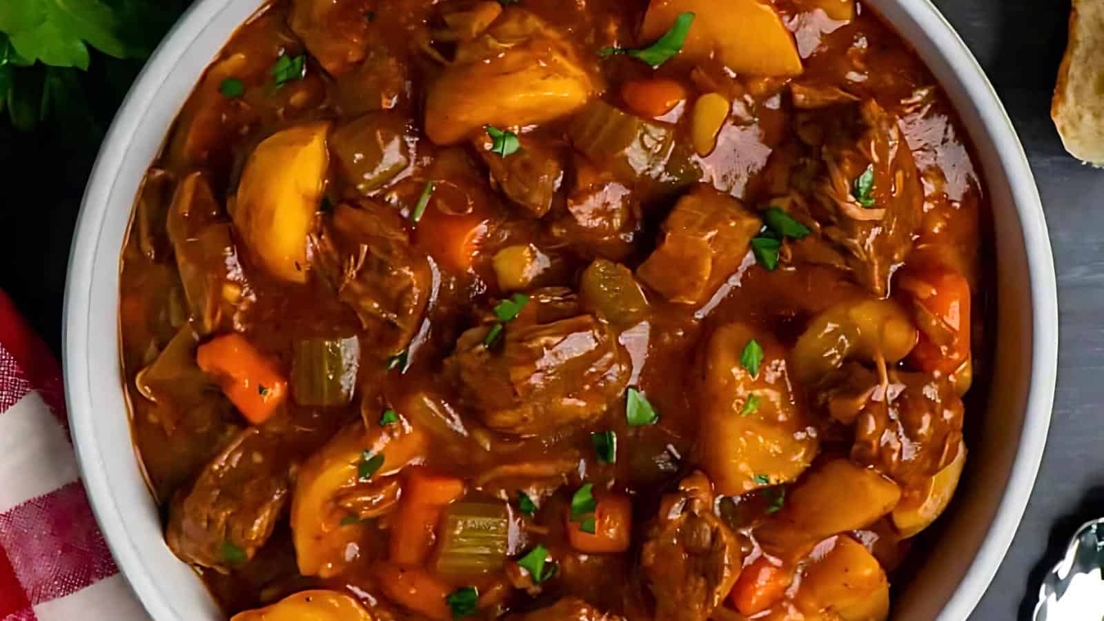 A bowl of hearty beef stew is filled with chunks of tender meat, diced potatoes, carrots, and celery, all in a rich, dark gravy. Garnished with chopped parsley, the dish looks flavorful and comforting. A partial view of a red checkered napkin is visible on the side.